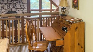 Un bureau en bois avec une lampe, une chaise et une bibliothèque, dans une pièce aux murs de pierre et à la rambarde en bois.