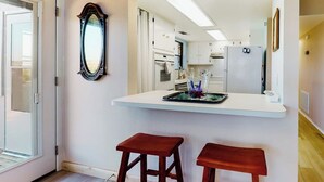 Une cuisine avec un plan de travail blanc, deux tabourets en bois, un miroir et un réfrigérateur.