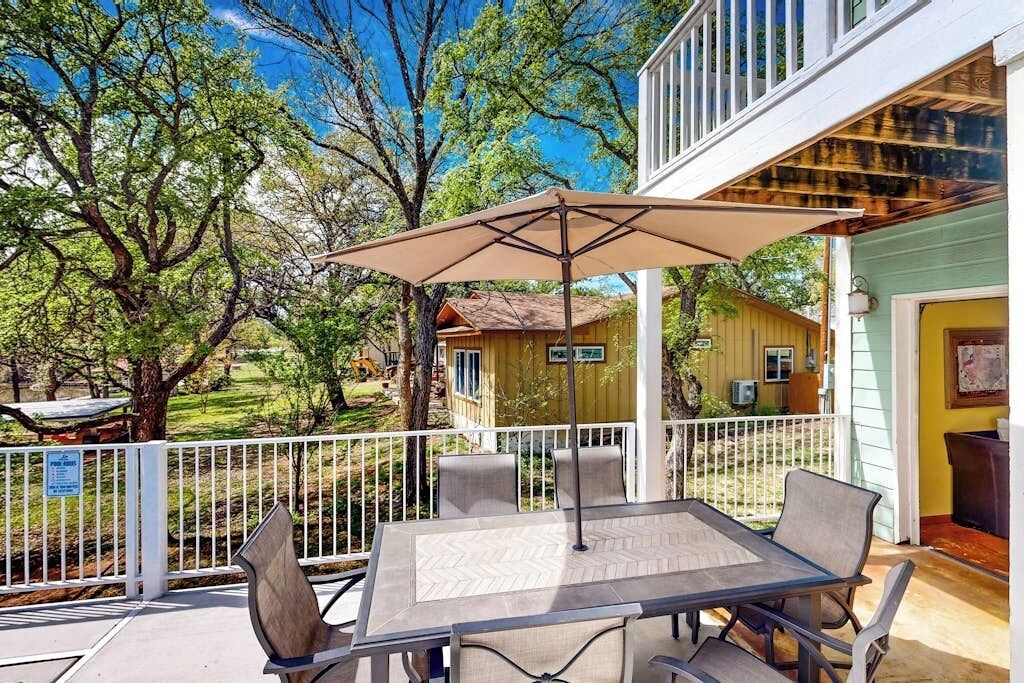 A covered patio with a table and chairs, surrounded by a white railing, overlooking a yard with trees and a small building.