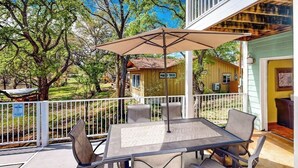 A covered patio with a table and chairs, surrounded by a white railing, overlooking a yard with trees and a small building.