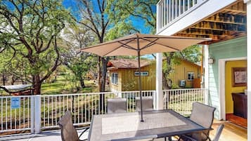 A covered patio with a table and chairs, surrounded by a white railing, overlooking a yard with trees and a small building.