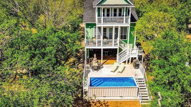 A two-story house with a balcony, a swimming pool, and a forested backdrop.