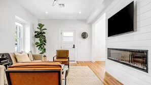 A modern living room with a fireplace, a large TV, and a plant. - Historic home a block from Main St with hot tub, sauna, fast WiFi & firepit (Fredericksburg)
