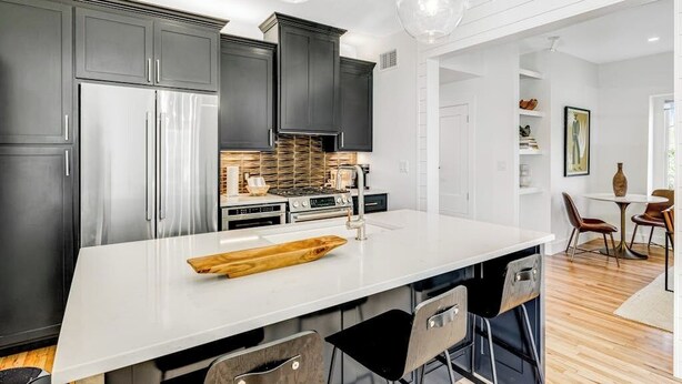 A modern kitchen with dark cabinets, stainless steel appliances, a white island, and a dining area with chairs and a table. - Historic home a block from Main St with hot tub, sauna, fast WiFi & firepit (Fredericksburg)