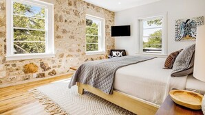 A bedroom with a stone wall, large windows, a bed, a ceiling fan, and a mounted TV. - Historic home a block from Main St with hot tub, sauna, fast WiFi & firepit (Fredericksburg)