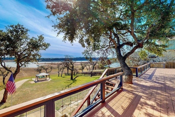 Une terrasse en bois, dotée d’une balustrade, qui donne sur une zone verdoyante avec des arbres et offre une vue lointaine sur une étendue d’eau.