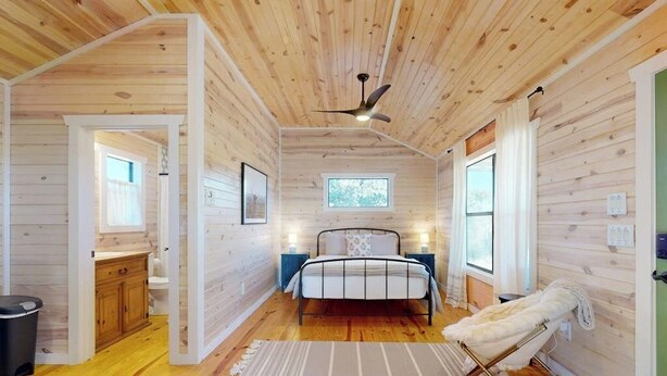 Interior de una cabaña de madera con una cama, lámparas de noche, un ventilador de techo y una ventana.