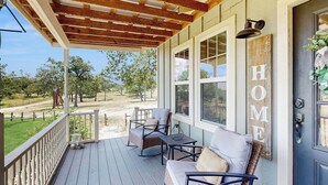 A covered porch with a wooden ceiling, a seating area with a sofa and chairs, large windows, and a 'HOME' sign on the door. - Cozy ranch home w/full kitchen, living room, hot tub, pet-friendly & fireplace (Fredericksburg)