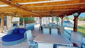 An outdoor patio with a stone fireplace, a round blue sofa, a table with chairs, and a bar area. - Spicewood Lake Retreat | Pool + Shuffleboard (Spicewood)