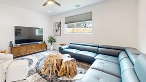 Un salon moderne avec un canapé d’angle, un téléviseur à écran plat, un ventilateur de plafond et une grande fenêtre.