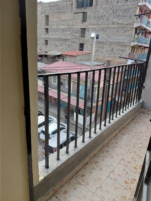 Ein Balkon mit Fliesenboden, Metallgeländer und Blick auf eine Straße mit Gebäuden und parkenden Autos.