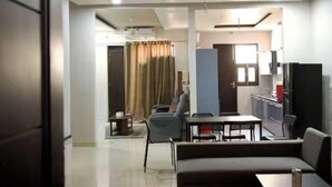 A living room with a sofa, a dining table, and chairs. There is a kitchen area with cabinets and a refrigerator. - Amur Stays 4: 6 Bedroom Independent Floors (Sahibzada Ajit Singh Nagar)
