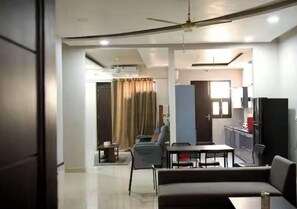 A living room with a sofa, a dining table, and chairs. There is a kitchen area with cabinets and a refrigerator.