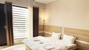 A hotel room with a bed, a nightstand, a lamp, and a window with curtains. - Amur Stays 4: 6 Bedroom Independent Floors (Sahibzada Ajit Singh Nagar)