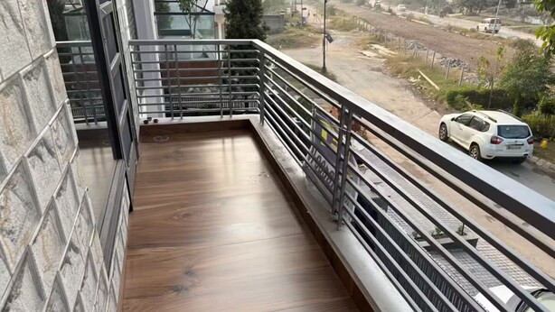 A balcony with a wooden floor and metal railing, overlooking a road with cars. - Amur Stays 4: 6 Bedroom Independent Floors (Sahibzada Ajit Singh Nagar)