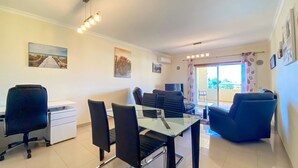 Uma sala de estar moderna com uma mesa de jantar de vidro, cadeiras pretas, uma escrivaninha branca e uma grande janela com vista para o verde.