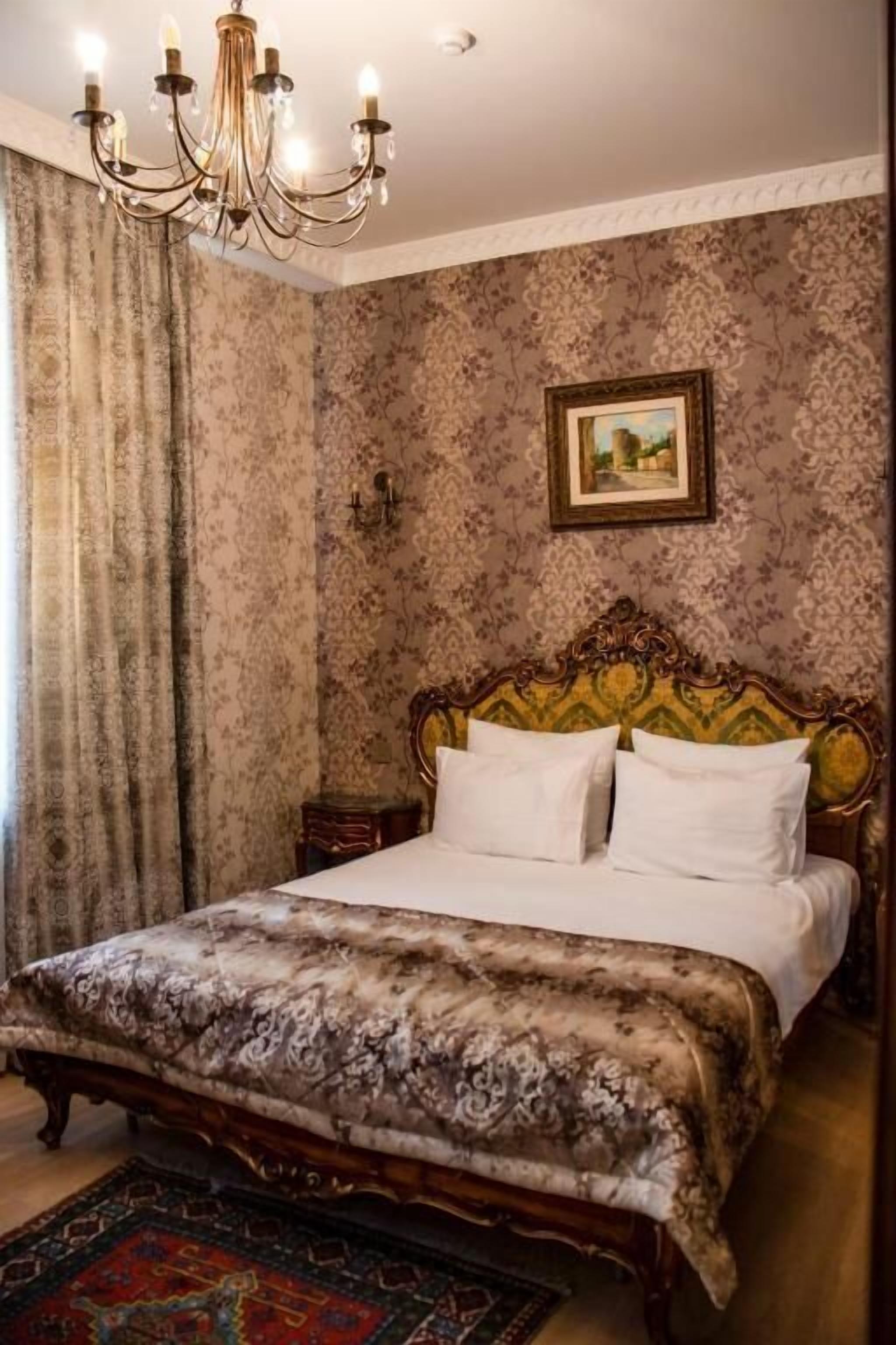 Un dormitorio con una cama grande, un cabecero ornamentado, una araña, papel tapiz estampado y un cuadro en la pared.