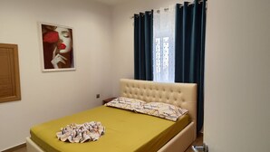 Un dormitorio con cama, ventana con cortinas, luz adosada a la pared y un cuadro.
