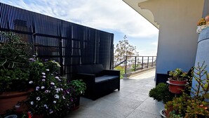 Un balcón con un sofá negro, plantas en macetas y un edificio moderno al fondo.
