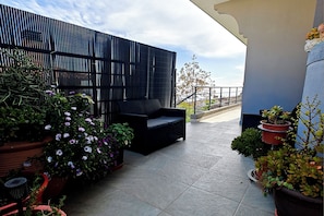 Un balcon avec un canapé noir, des plantes en pot et, en arrière-plan, un immeuble moderne.