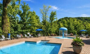 Ein Schwimmbecken mit Liegestühlen und Sonnenschirmen, umgeben von Bäumen und einer hügeligen Landschaft.