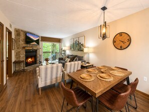 Ein gemütliches Wohnzimmer mit Kamin, eine Essecke mit Holztisch und gewebten Untersetzern und eine große Wanduhr.