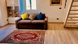 Un salon avec un canapé marron, un tapis à motifs, une fenêtre avec des stores, un tableau encadré au mur et un meuble blanc avec des portes en verre.