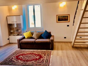 Ein Wohnzimmer mit einem braunen Sofa, einem gemusterten Teppich, einem Fenster mit Jalousien, einem gerahmten Bild an der Wand und einer weißen Kommode mit Glastüren.