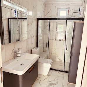 A modern bathroom with a marble backsplash, a sink, a toilet, a shower enclosure, and a refrigerator.