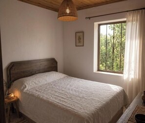 A bedroom with a wooden headboard, a bedside table with a lamp, a window with curtains, and a framed picture on the wall.