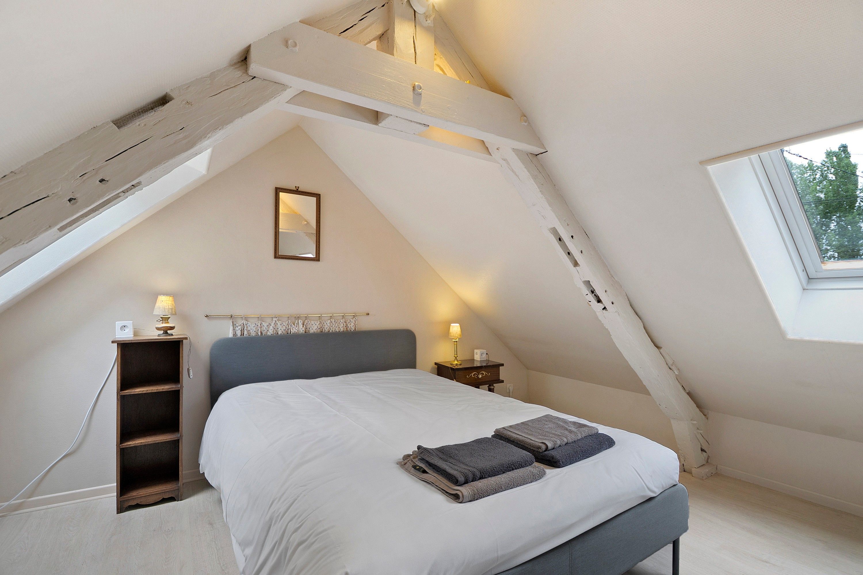 Une chambre avec un plafond en pente, un lit avec une tête de lit grise, des lampes de chevet, une petite table de chevet, un miroir fixé au mur et une fenêtre donnant sur des arbres.