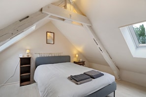 A bedroom with a sloped ceiling, a bed with a grey headboard, bedside lamps, a small bedside table, a wall-mounted mirror, and a window with a view of trees.
