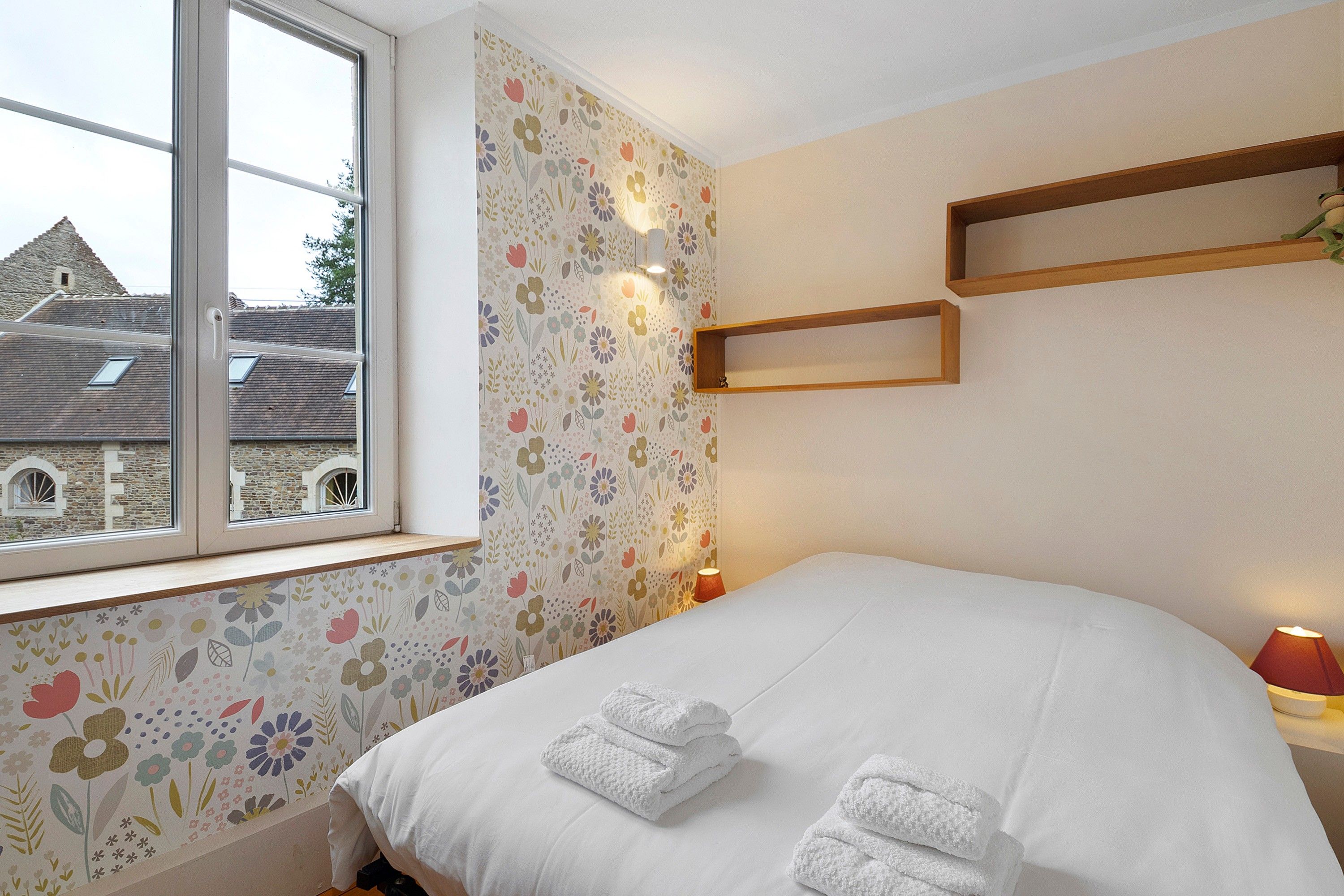 A bedroom with a bed, a window, a floral wallpaper, and a view of a stone building.