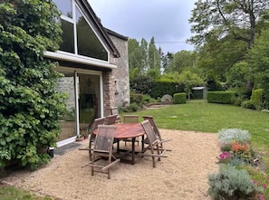 A patio with a table and chairs, a stone building with a glass door, and a well-maintained garden with hedges and flowers.