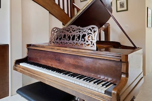 A grand piano with a wooden finish and a decorative stand in a room with a staircase and framed pictures on the wall.