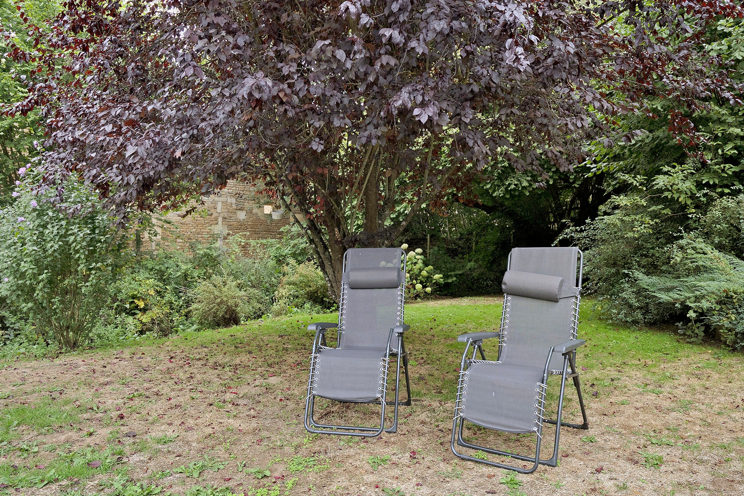 Deux fauteuils de jardin, avec une structure en métal et un assise en tissu, sont placés sur une pelouse, avec en arrière-plan un feuillage dense et un mur de pierre.