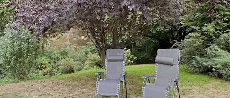 Deux fauteuils de jardin, avec une structure en métal et un assise en tissu, sont placés sur une pelouse, avec en arrière-plan un feuillage dense et un mur de pierre.