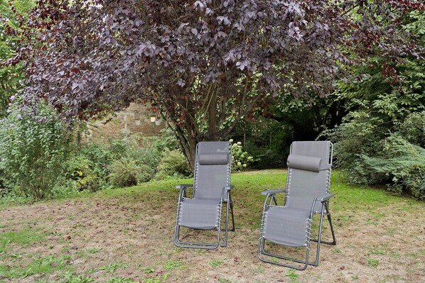 Two outdoor lounge chairs with metal frames and fabric seats, positioned on a grassy area with a backdrop of dense foliage and a stone wall.