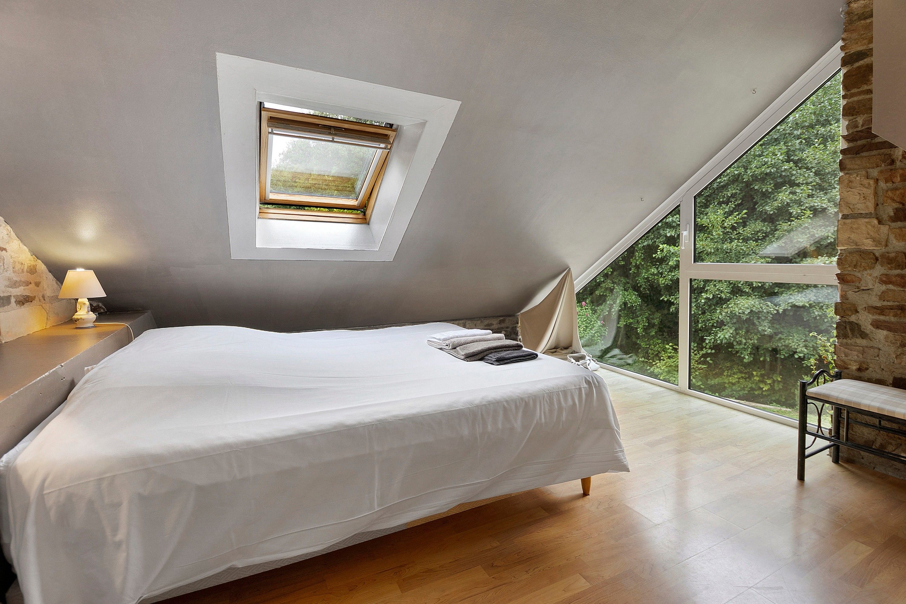 Une chambre avec un plafond en pente, un lit, une table de chevet, une lampe et une grande fenêtre donnant sur un paysage verdoyant.