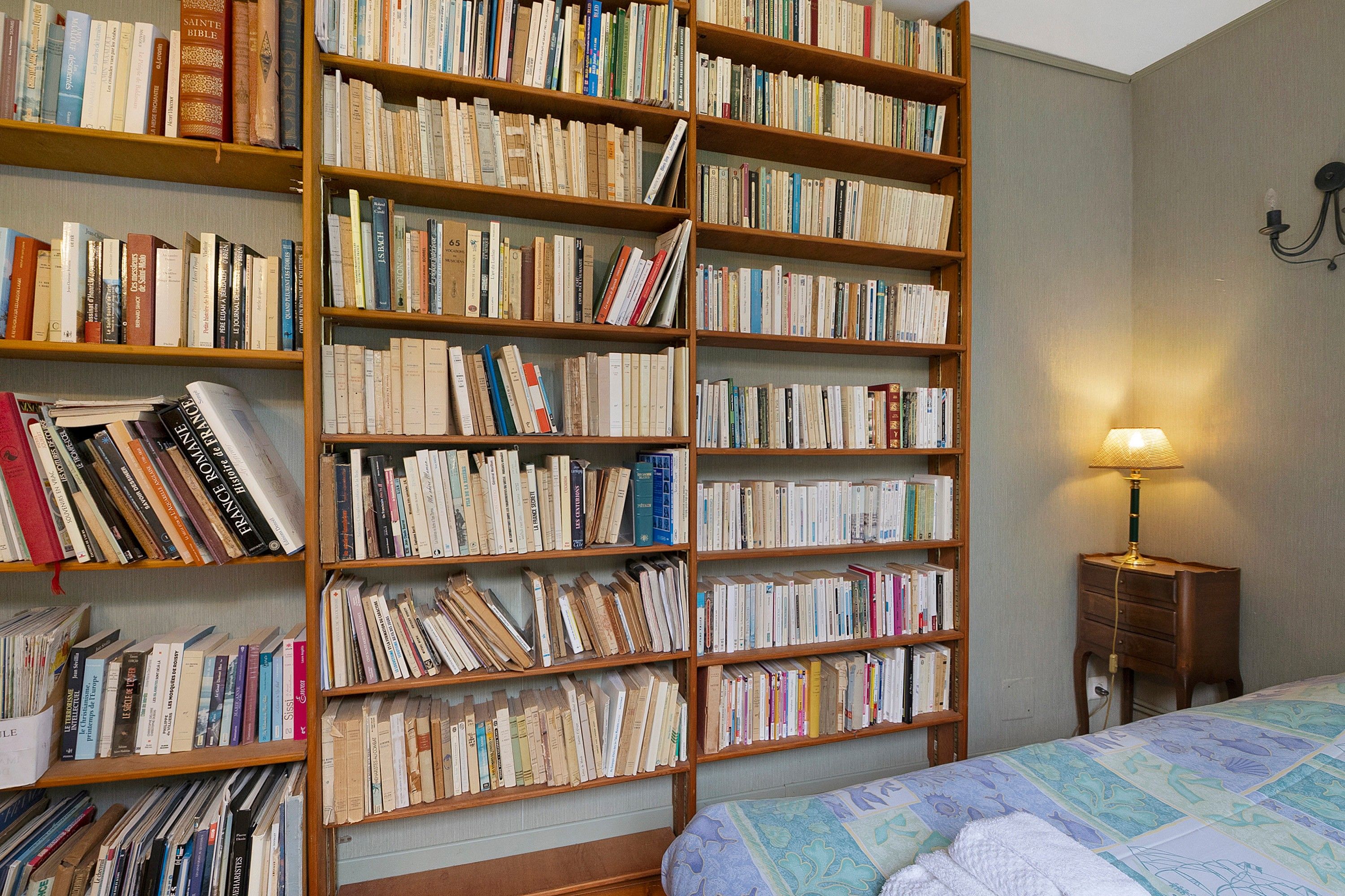 A room with a large bookshelf filled with books, a bed with a patterned bedspread, and a bedside lamp.