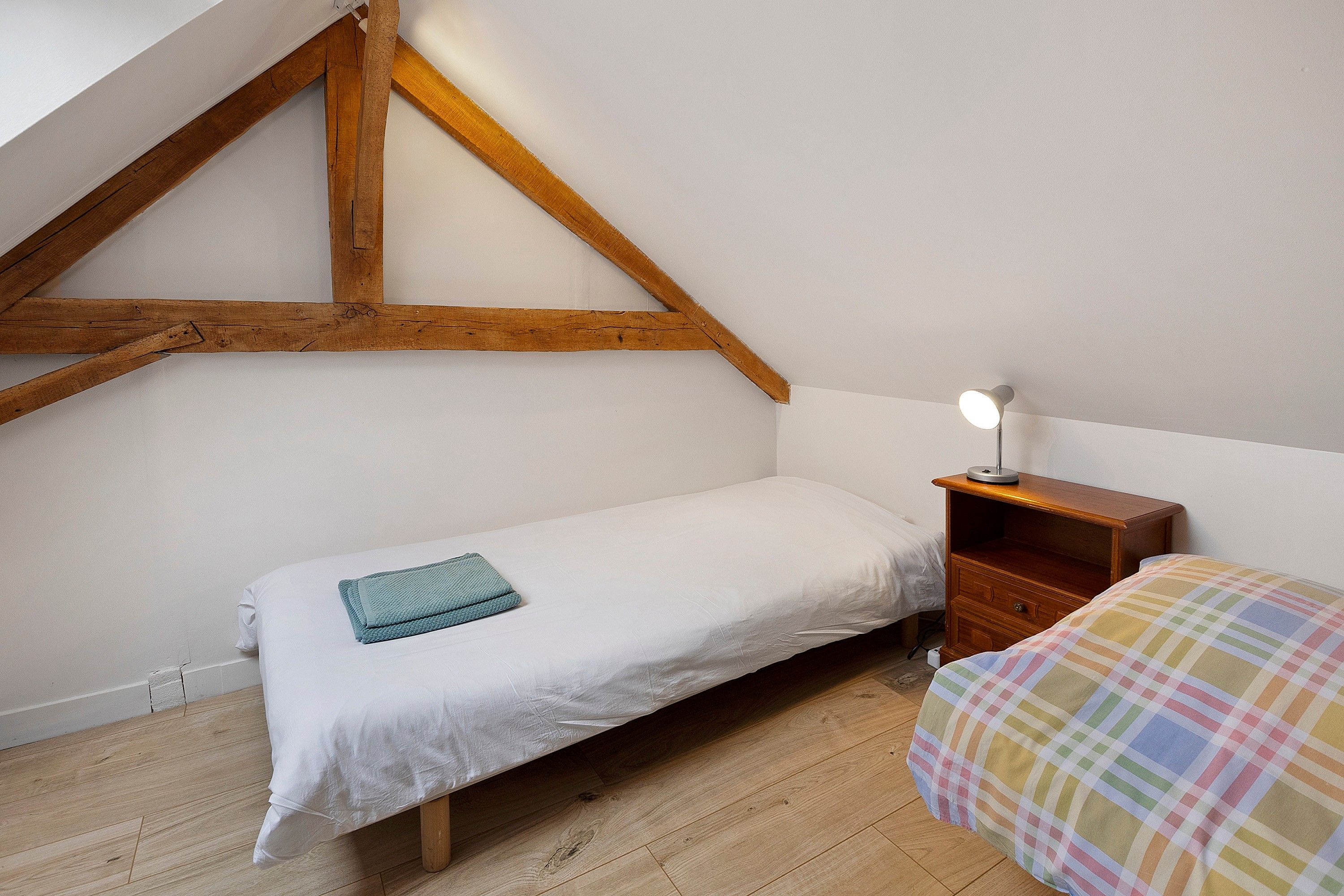 Ein kleines Zimmer mit einem Einzelbett, geneigter Decke und Holzbalken, einem Nachttisch mit Lampe sowie einer farbenfrohen, karierten Bettdecke.