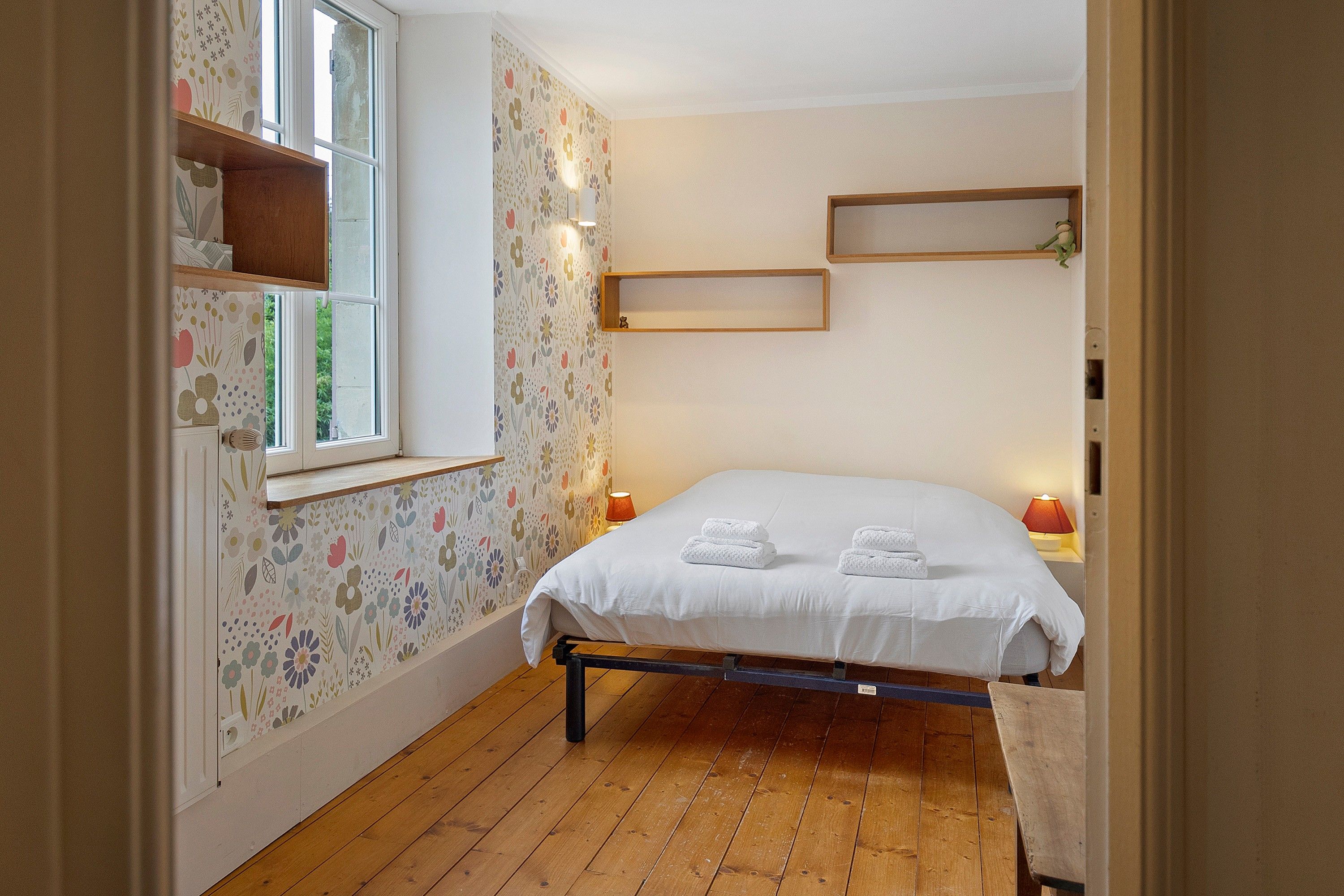 Une chambre à coucher avec un lit, un plancher en bois, un papier peint à motifs floraux et une fenêtre.