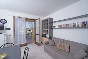 Uma sala moderna com um sofá cinza, uma estante de livros, um espaço de refeições com mesa e cadeiras, e um painel de vidro deslizante que dá para uma área externa.