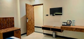 Una habitación de hotel con un banco de madera, un televisor montado en la pared, una cafetera y un cubo de basura.