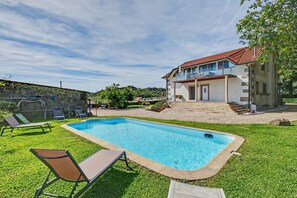 A swimming pool with lounge chairs, a two-story house with a balcony, and a stone wall in the background.