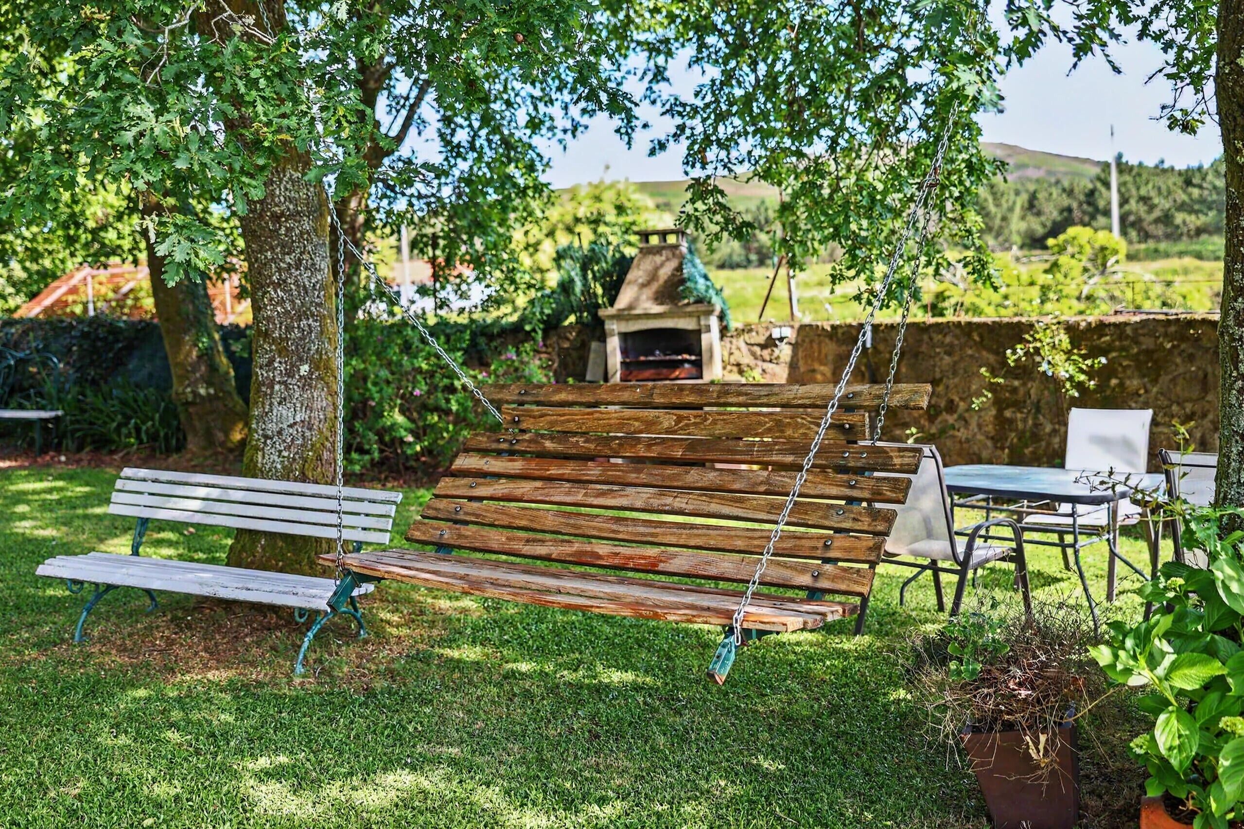 Un banc-balançoire en bois sous un arbre, un coin salon extérieur rustique avec un mur en pierre et un petit foyer au fond.