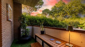 Un banco de madera, una mesa y una planta en maceta en un rincón, junto a una pared de ladrillo y vistas a árboles.