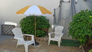 Deux chaises en plastique blanches, un parasol jaune et blanc, et des plantes vertes dans un espace patio.