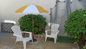Zwei weiße Plastikstühle, ein gelb-weißer Sonnenschirm und grüne Pflanzen auf einer Terrasse.