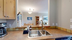 Una cocina con un lavamanos doble, armarios de madera, una cafetera, una estantería y un comedor con sillas.